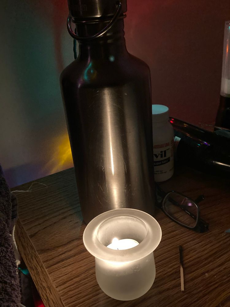 A lit candle near edge of wood topped bedside table, behind candle is metal water bottle, pair of glasses, bottle of advil, burnt match next to candle. Candle reflects on white background wall and there are coloured lights visible at the edges of the photo.