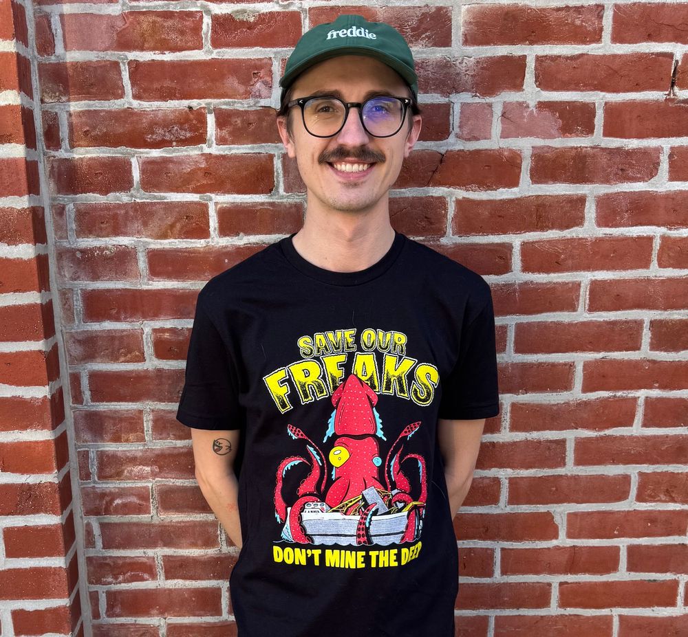 My grad school roommate nick, who is an extremely adorable bespeckled and mustacio'd Canadian twink, stands against a brick wall wearing a black t-shirt with the words save our freaks don't mine the deep, with a big strawberry squid sinking a mining ship