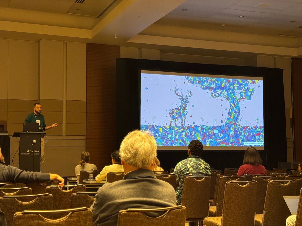 Kevin kohl, a tall handsome man in a green sweater, talks with his hands in front of a pretty slide of a stag and tree artistically overlaid with microbes in many colors 
