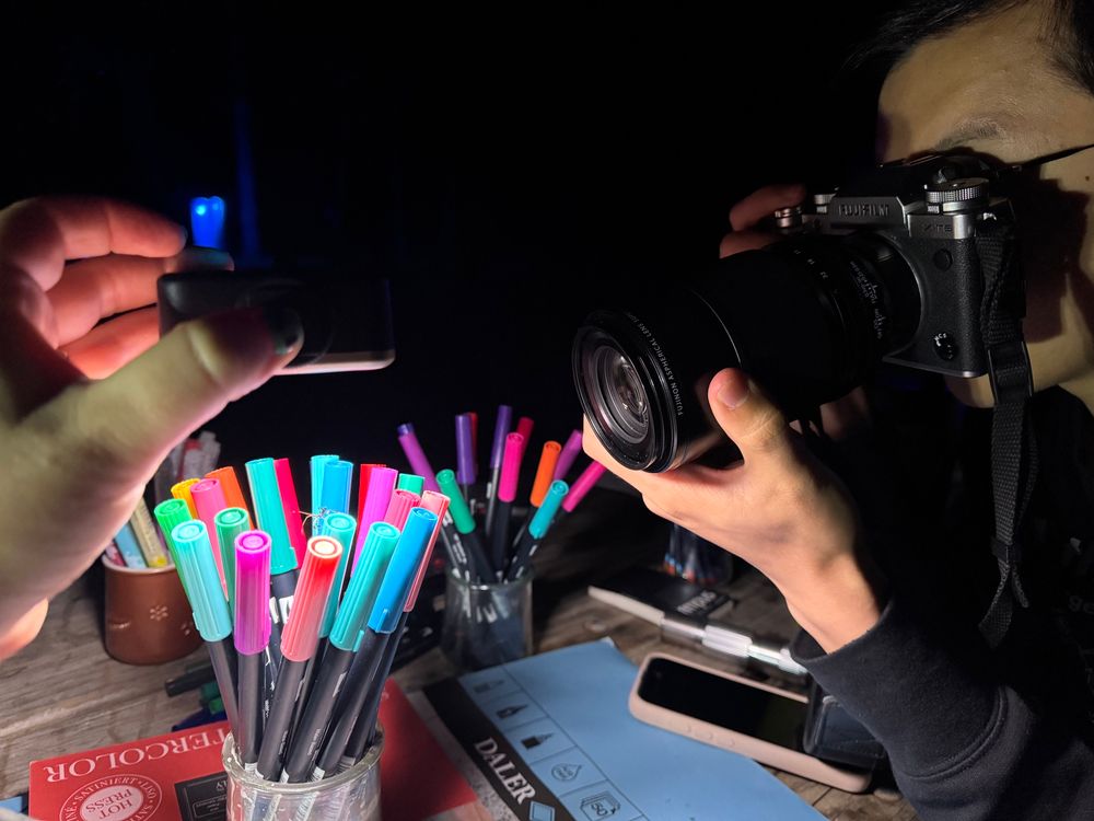 Sarah holds up a light box over a jar of colorful markers with Sean on the camera taking a picture of a tiny little bug who is standing on the colorful marker caps