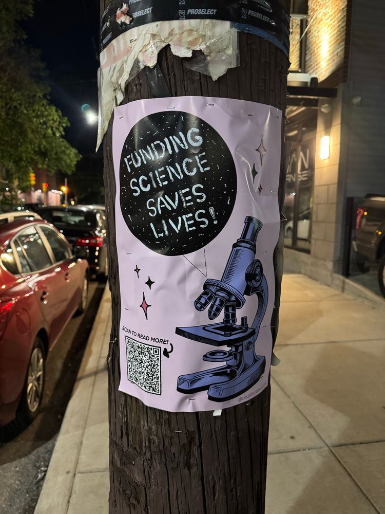 A poster with a microscope shows a field of microbes that have all arranged themselves to say “funding science saves lives” and a big ol QR that says “scan to learn more”, stapled to a telephone pole on a city street