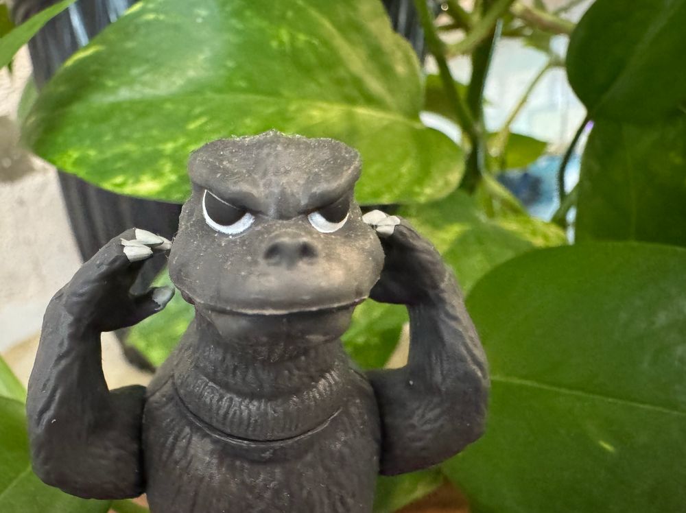 a small godzilla toy stands in front of a pothos plant with his claws by his face with angry eyes. He is frustrated!!!! but he only has himself to blame!!!!