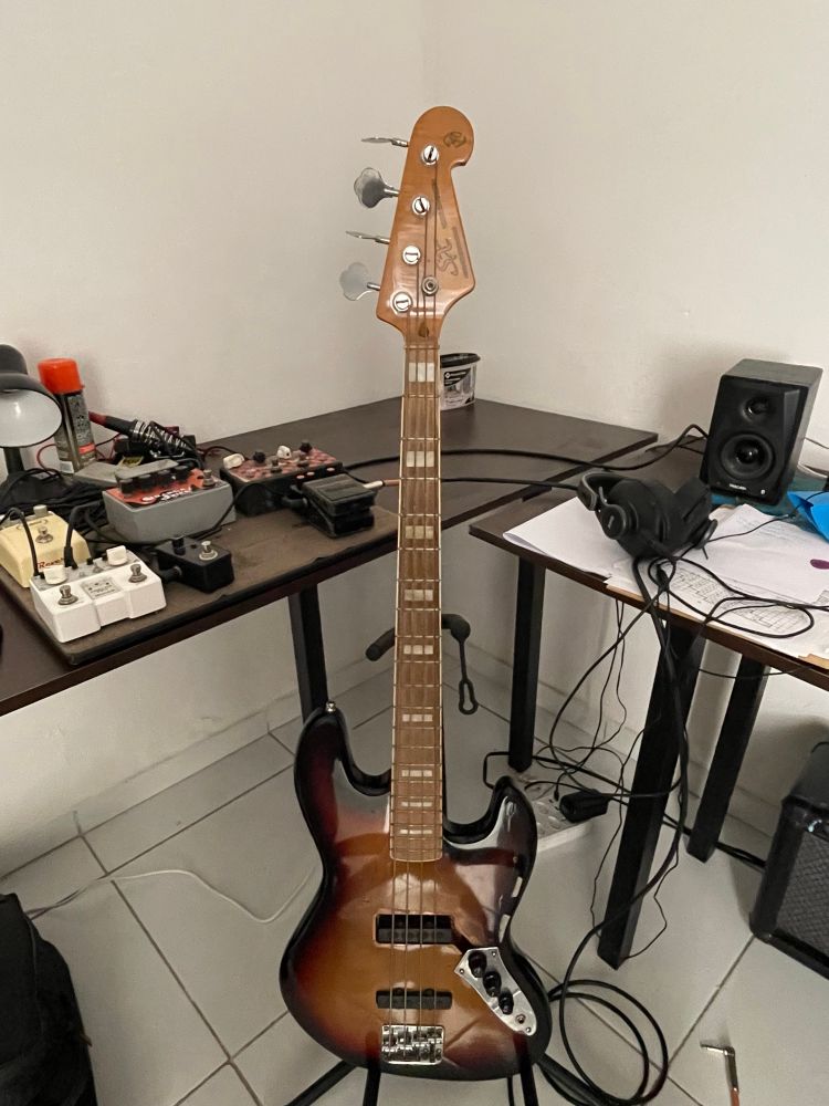 Foto de um contrabaixo jazz bass, ao fundo sobre uma bancada pedais de guitarra, fones de ouvido, cabos entre outras coisas 