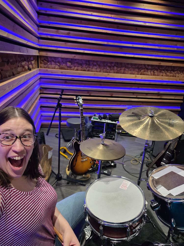 A selfie of me smiling inside a music studio with a drum kit in the front, a guitar behind it, and wood walls with blue Phillips Hue lightstrups
