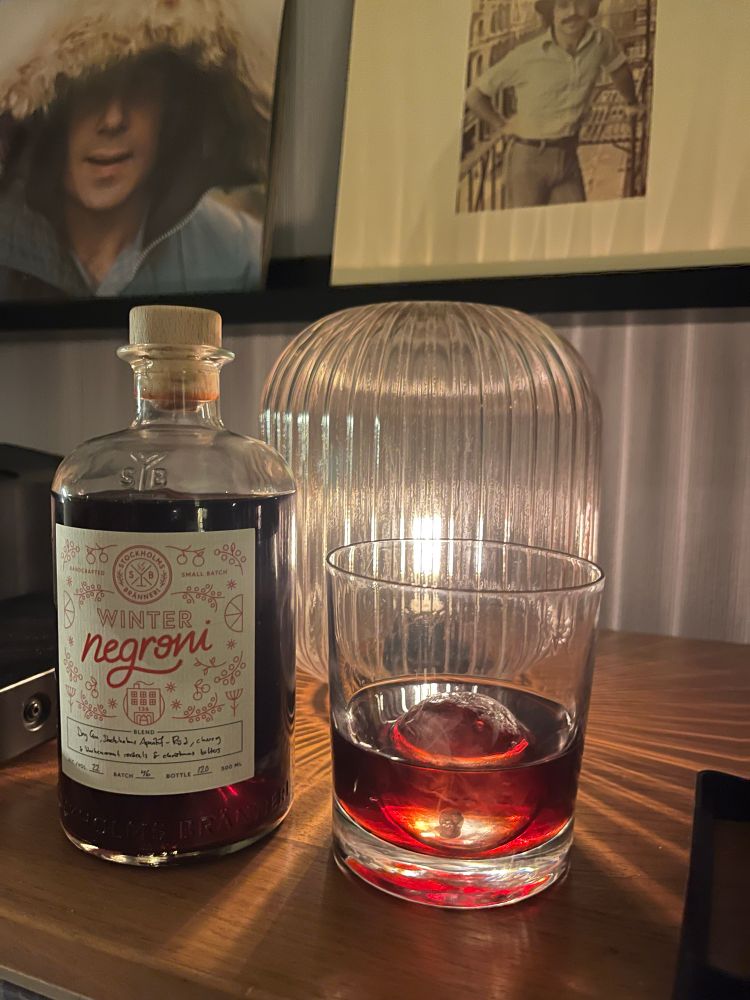 En flaska Winter Negroni och ett glas på ett bord. En lampa lyser bakom och lyser upp den röda vätskan