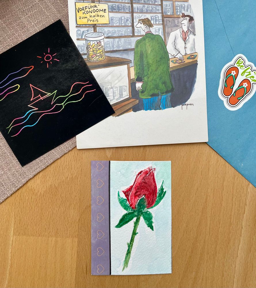 Auf einer hölzernen Tischplatte liegend: mittig ein ATC Kärtchen bemalt mit eine Rose plus Stiel, darüber liegen von links qnach rechts ein quadratisches kleines schwarzes Bild mit buntem Boot auf Wellen und Sonne,  eine Postkarte die zwei Männer an einem Tresen zeigen und ein blauer Briefumschlag mit einem Aufkleber in Form von Flip Flops