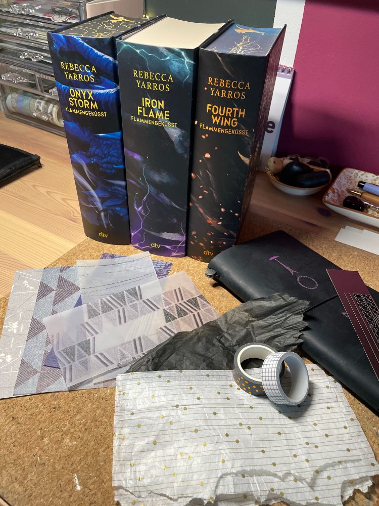 Drei dicke gebundene Bücher mit buntem Einband stehend auf einem Schreibtisch. Im Vordergrund unterschiedliche Papierreste, Washitape und ein Notizbuch. Im Hintergrund Bastelmaterialien. 