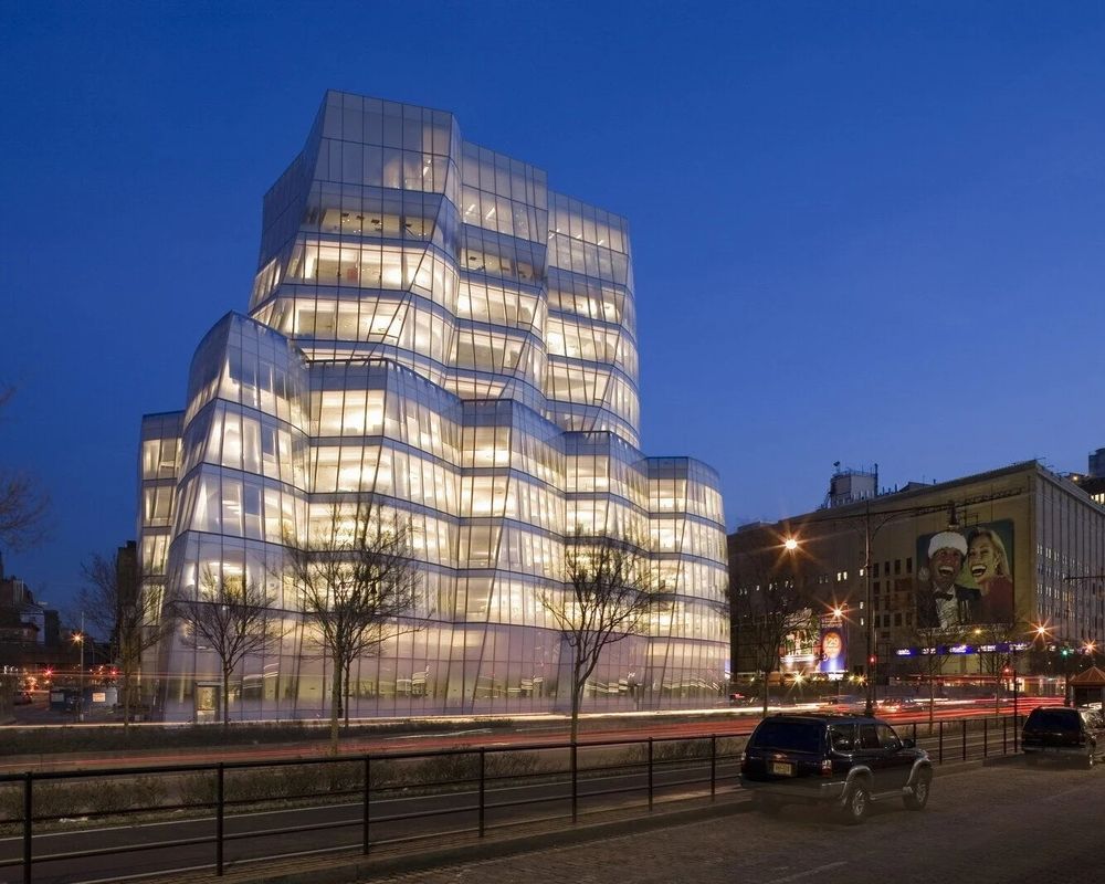 The Frank Gehry-designed IAC Building in Manhattan. The building is lit up and is set against a late dusk sky. In the foreground is a roadway, some small winter-bare trees, and two parked vehicles.