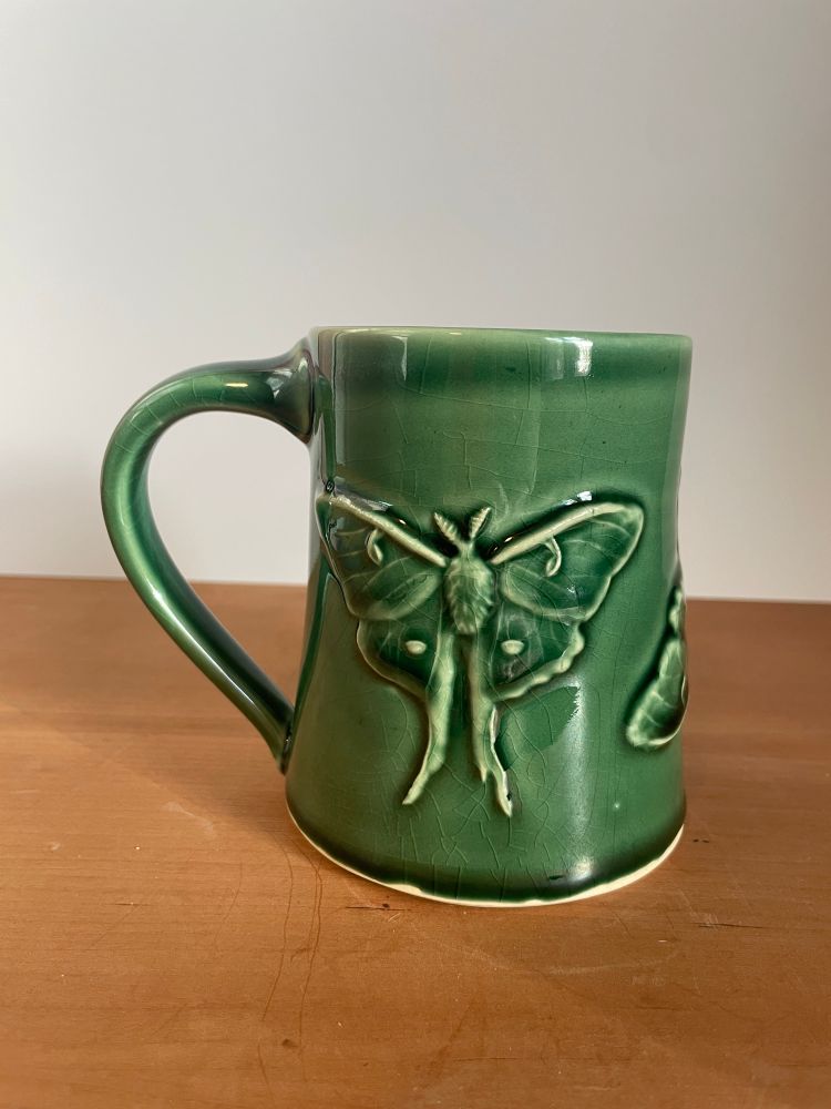 An emerald green translucent glazed mug, the mug has a bas relief of a Luna moth, the glaze is thin on high spots and thick in low spots creating further depth