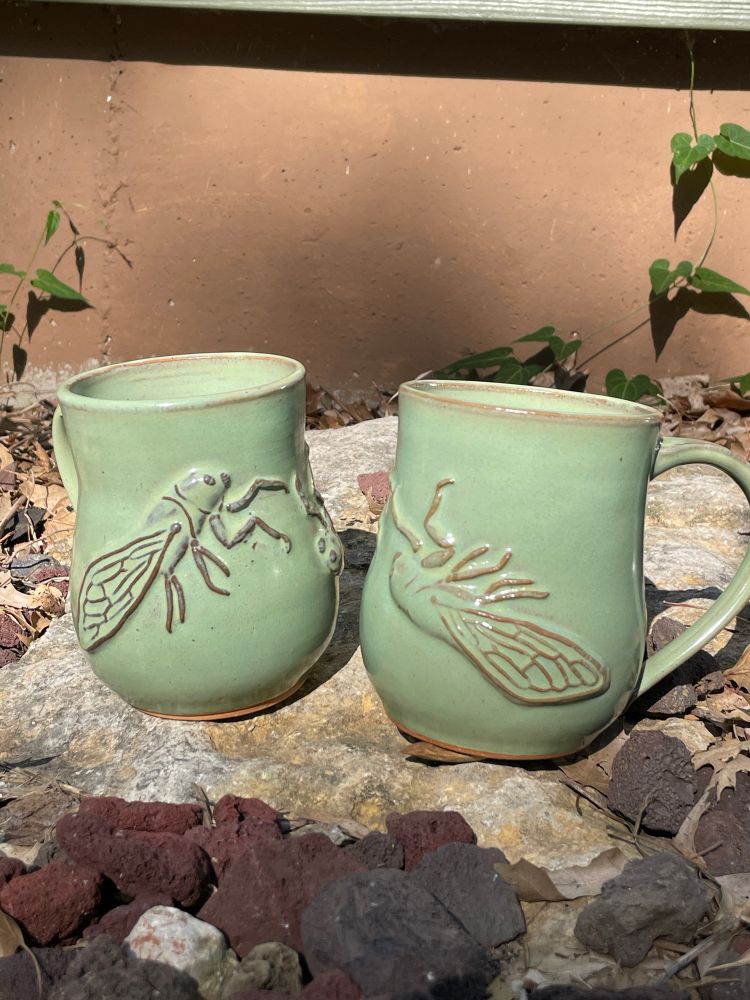 Two light green mugs with bas relief cicadas sit on a rock