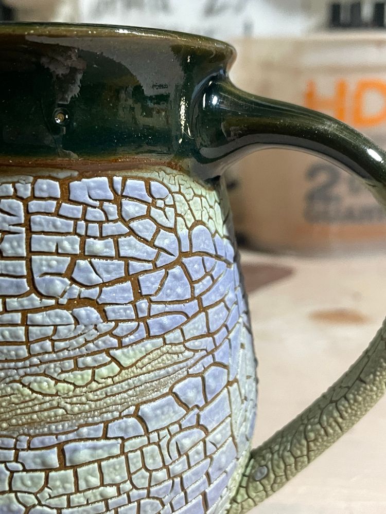 Detail at the handle joint, A coffee mug with a lower section of crackled light green/blue glaze that looks dried earth, a dark green glaze is at the top