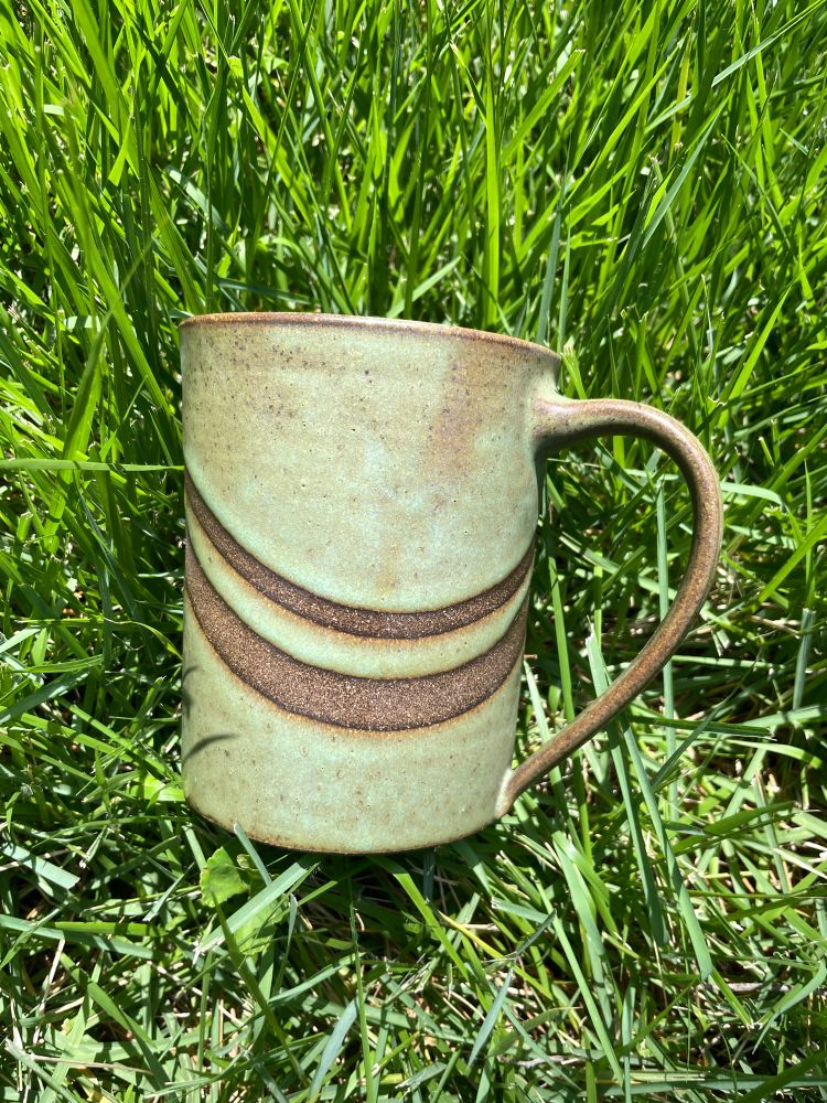 A light green mug with dark brown bands sits on bright green grass 