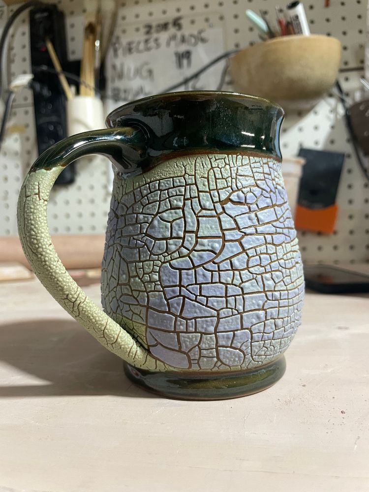 A coffee mug with a lower section of crackled light green/blue glaze that looks dried earth, a dark green glaze is at the top