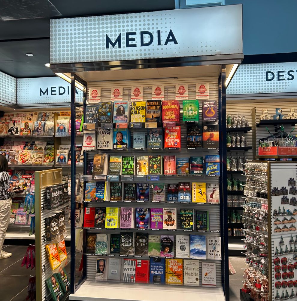 Shelf at an airport store a la Hudson news entirely stocked with books and above the shelf it says MEDIA