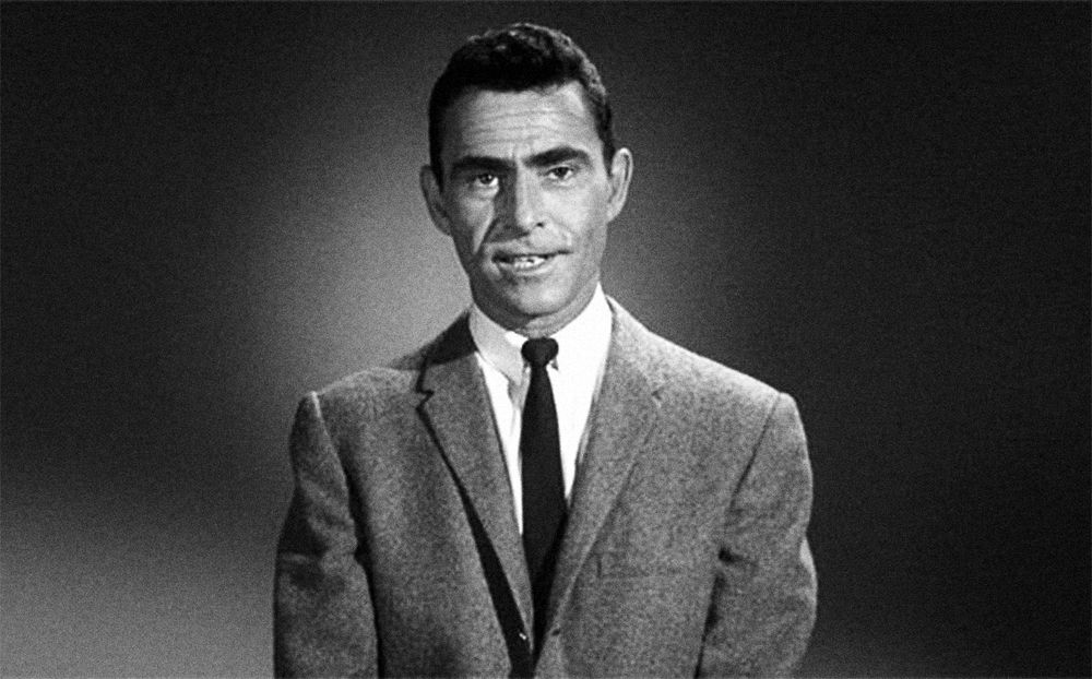 Rod Serling, middle aged white man, host of the twilight zone, addressing camera in one of his signature show opening monologues. He has short black hair and a grey suit jacket and black tie, the background is a neutral grey, black and white image