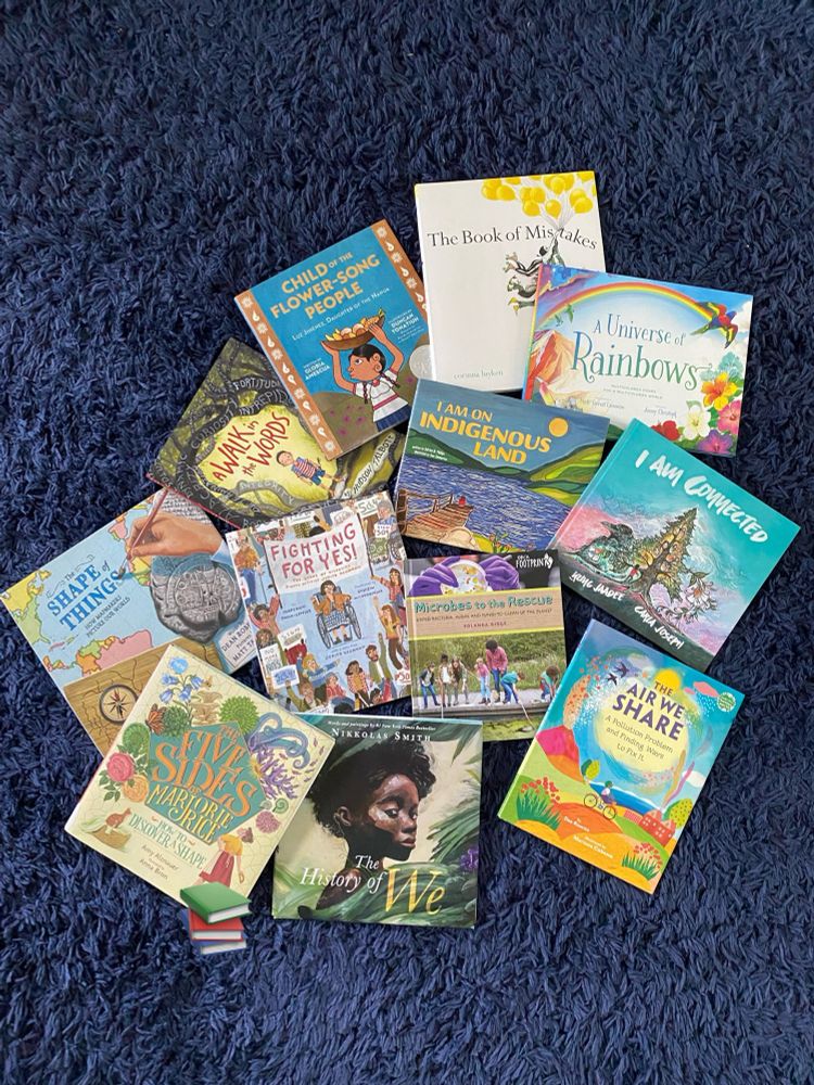 Books scattered include Shape of Things, A Walk in the Words, Child of the Flower Song People, The Book of Mistakes, A Universe of Rainbows, I am on Indigenous Land, I am Connected, Fighting for Yes, Microbes to the Rescue, The Air We Share, The History of We, and The Five Sides of Marjorie Rice
