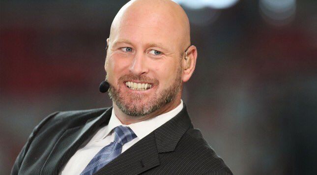 Trent Dilfer smiling on the sideline of a football game, wearing a suit and a broadcaster headset