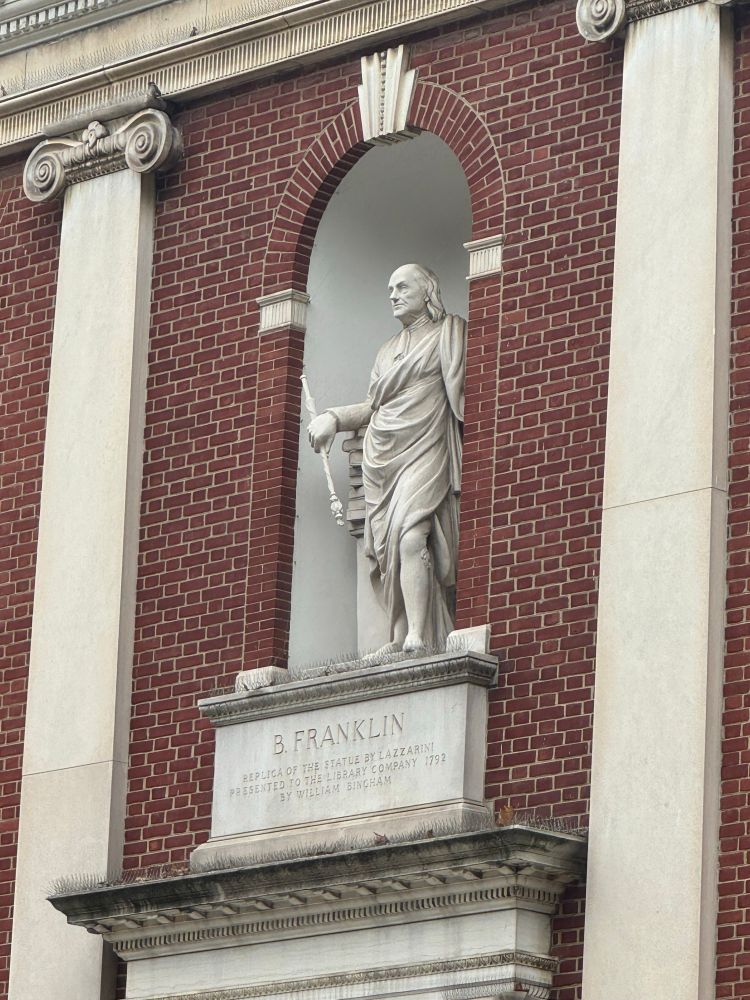 White marble statue of Benjamin Franklin in classical robes, holding a scepter, set within an arched niche on a red brick building facade. The plinth reads 'B. FRANKLIN'