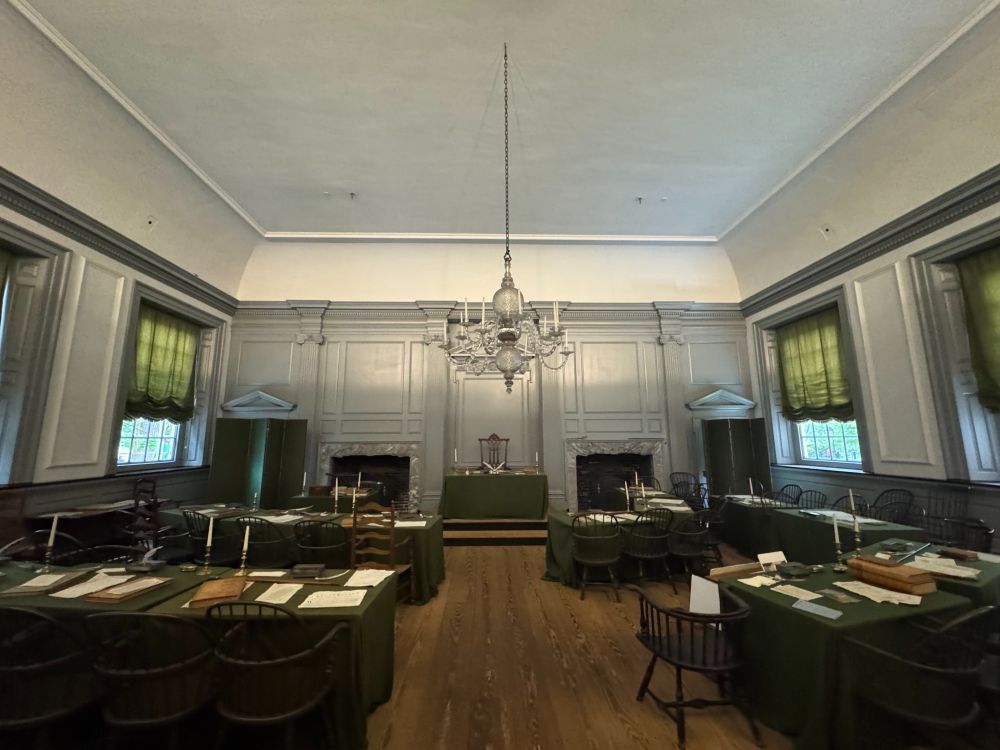 Interior of Independence Hall in Philadelphia, showing the Assembly Room where the Declaration of Independence and U.S. Constitution were debated. The room features green-covered tables, wooden chairs, tall windows with green drapes, two fireplaces, and a central chandelier.