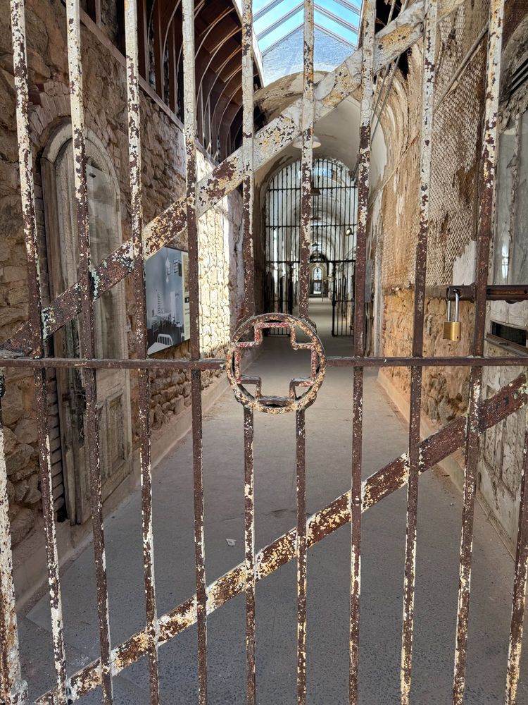 Rusty iron gate with peeling paint blocks entry to a long, narrow hallway in the hospital wing of Eastern State Penitentiary. The corridor beyond is lined with decaying stone walls, barred doors, and a vaulted skylight that lets in shafts of daylight. A padlock secures the gate, and layers of age and neglect are visible throughout the scene.