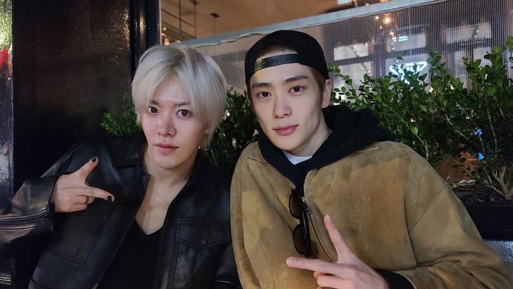 Yuta and Jaehyun sitting beside each other in front of some shrubbery. They are leaning in towards each other and are each making a peace sign with one hand. 