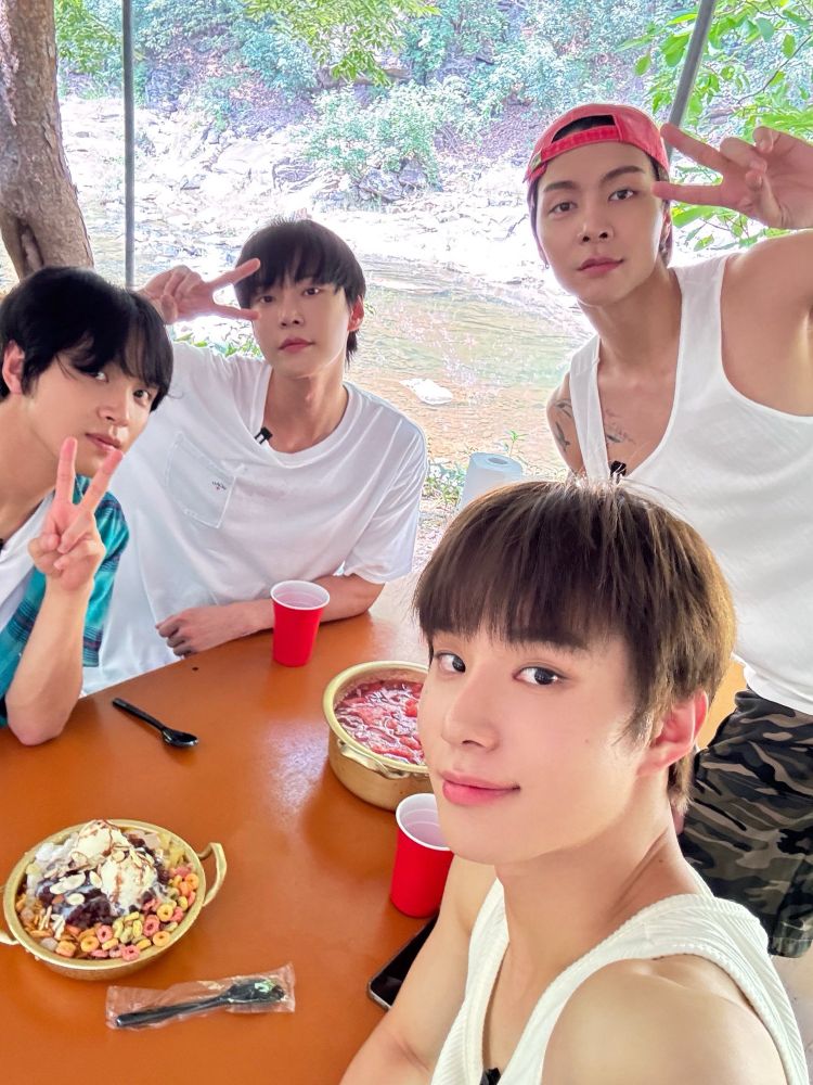 Haechan, Doyoung, Johnny, and Jungwoo sitting at a table next to a stream eating dessert while filming an NCT 127 Vibe content.