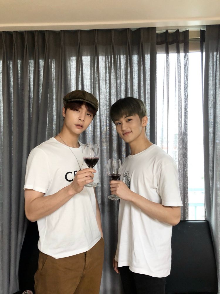 Johnny and Mark wearing casual clothes, holding wine glasses with a bit of red wine/grape juice in them, standing in front of a large window in their dorm after filming a VLIVE during the early months of the COVID pandemic.