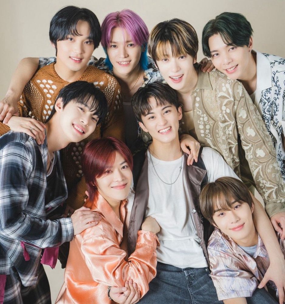 Doyoung, Haechan, Taeyong, Jaehyun, Johnny, Yuta, and Jungwoo around closely huddled around Mark who is sitting in a chair. They all have their arms around each other and are smiling.