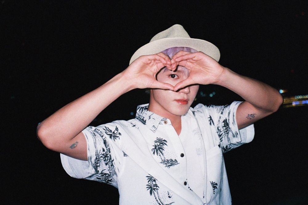 Taeyong standing outside at night. He’s wearing a beige fedora and a white button down shirt with black palm trees. He’s making a heart with his hands in front of one eye. 