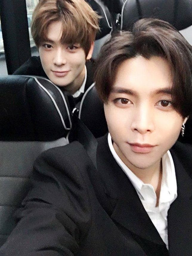 A selfie taken by Johnny with Jaehyun. They are sitting on a bus. Johnny is sitting in the aisle seat. Jaehyun is sitting in the window seat in the row behind Johnny. Jaehyun is leaning forward in his seat to be closer to the camera. 