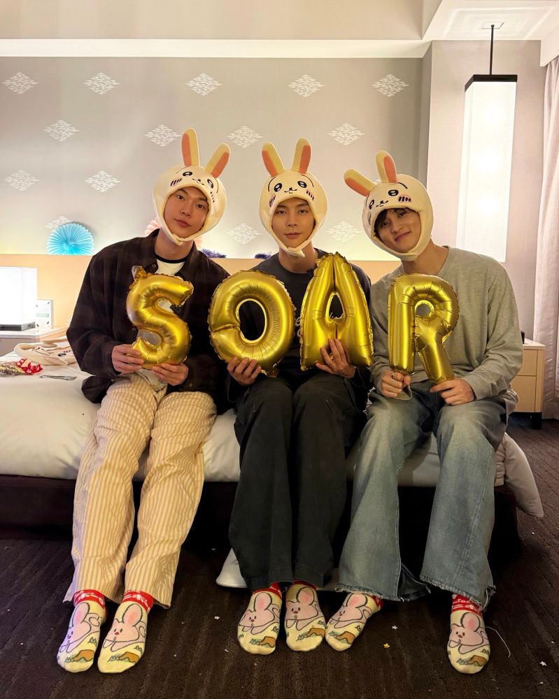 Doyoung, Johnny, and Mark sitting on a hotel bed wearing bunny ears and holding letter balloons that spell out SOAR during a listening party for Doyoung’s album. 