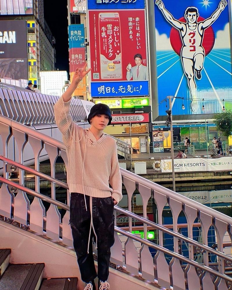 Taeyong standing on a set of stairs in Osaka at night in front of the iconic Glico billboard ad of a man running on a track. 