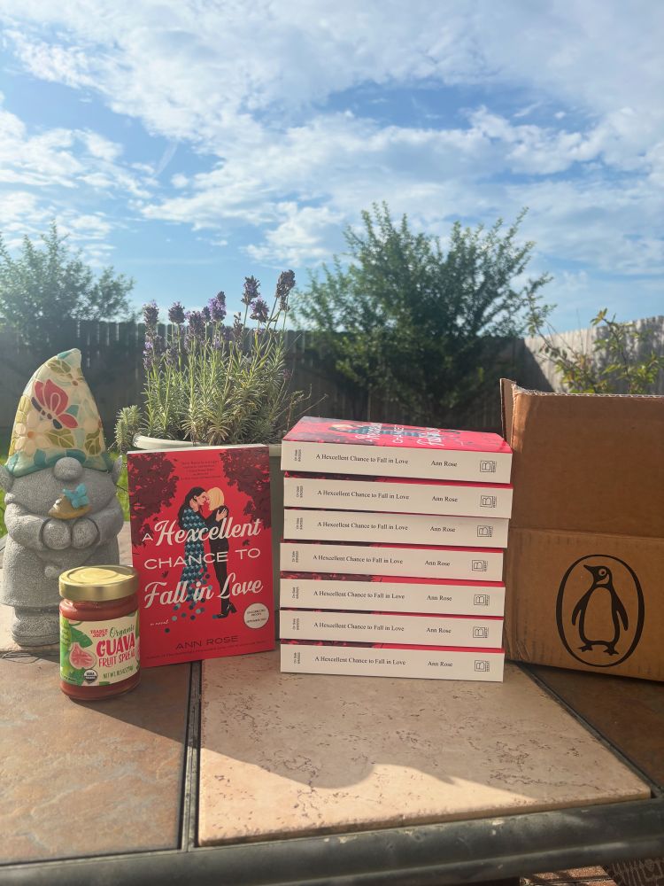 A bright blue sky in the background. A stack of books—A Hexcellent Chance to Fall in Love—sit on a table with a pot of lavender, a box from Penguin Random House, a gnome, and a jar of guava fruit spread from Trader Joe’s. The book cover has the image of two women on it, one slowly disappearing. 