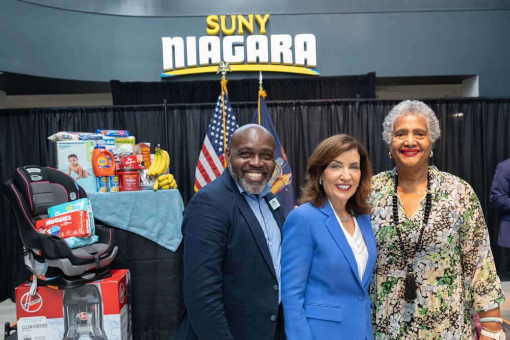 Governor Kathy Hochul poses with Trustee Lewin and President Holmes in front of a table filled with children's goods. A SUNY Niagara sign hangs in the background. 
