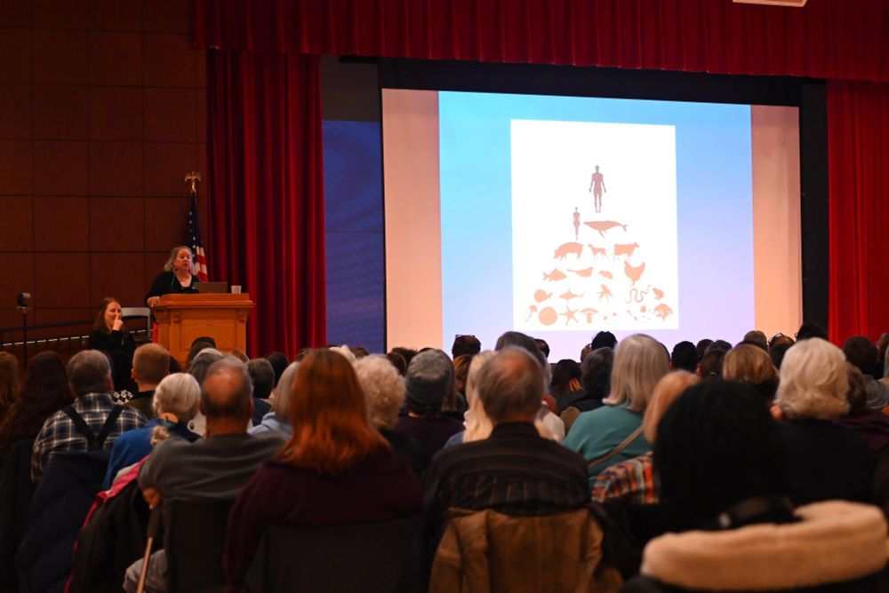 Dr. Robin Wall Kimmerer speaks on stage to a crowded auditorium with a display of images at SUNY Oneonta. 