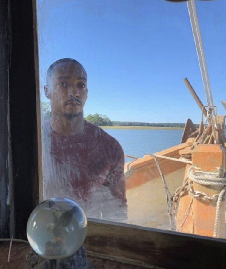 Anthony Mackie standing on the deck of a boat. He is photographed through the glass of the bridge. It's a beautiful sunny day, but he looks so sad. Please let him in. 