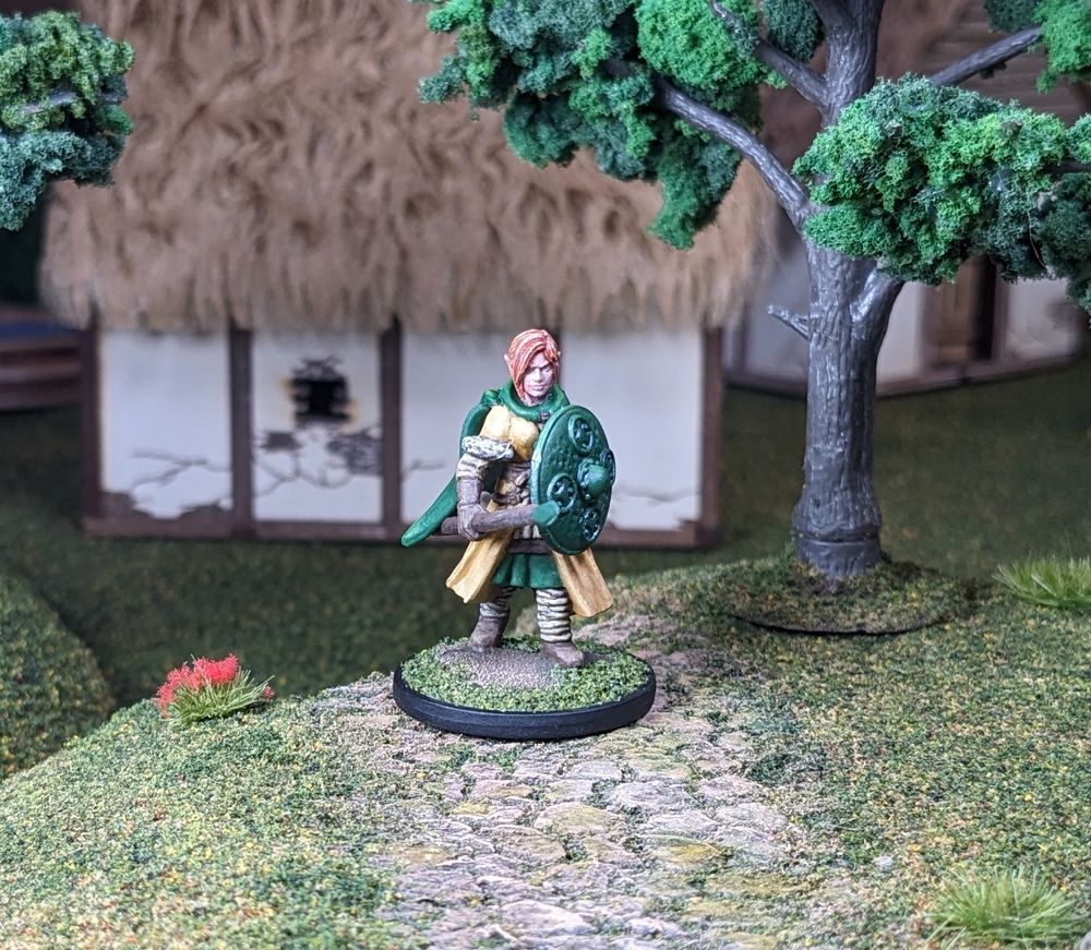 A red-haired female elf fantasy fighter miniature, armed with a spear and green shield. She is wearing a long beige coat with fur trim, and a green cloak. She is wearing sensible shoes, and clothing. She is facing forward.
