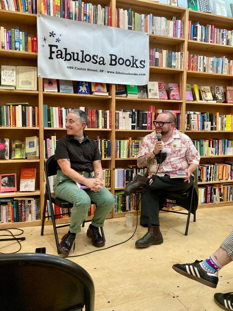 Quill and Elliot Kukla sit on foldable chairs in front of bookshelves and a banner reading "Fabulosa Books"