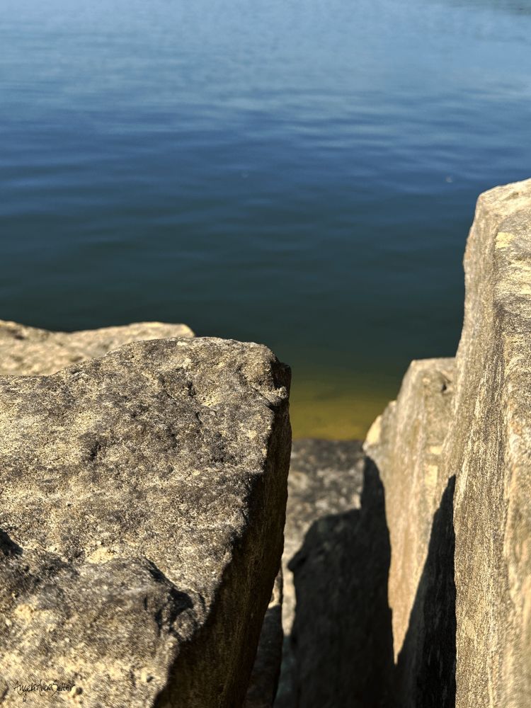 Large, square rock formation next to a lake. The stones are rough and the water is a smooth deep blue fading to a greenish hue at the shallow edge.