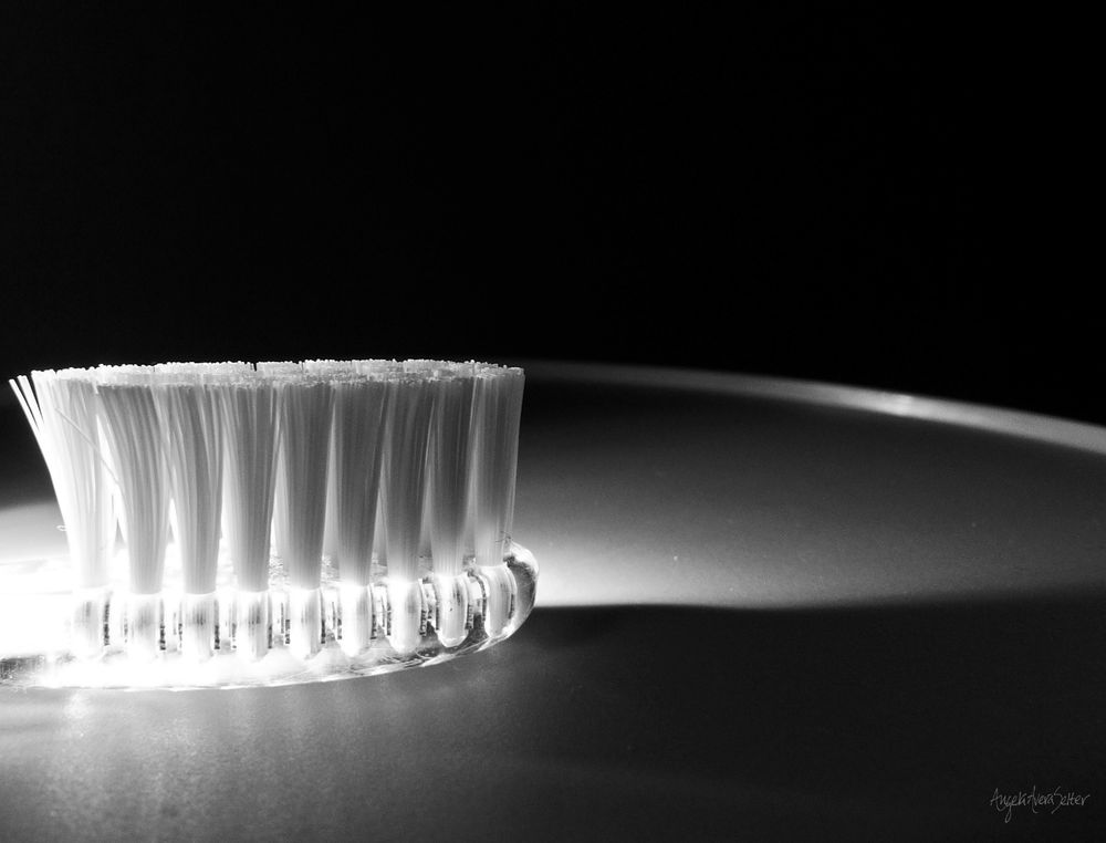 Head of a toothbrush. White bristles of even length set in a clear handle. It's resting on a smooth countertop tray and bright light is entering the frame from the left bleaching out the first group of bristles, then shading to grey as it's diffused toward the right side of the brush. The toothbrush is creating a shadow and light has captured the round edge of the tray in a smooth arced line which splits the image horizontally. The background beyond the tray edge is deep black and makes up the top third of the image.