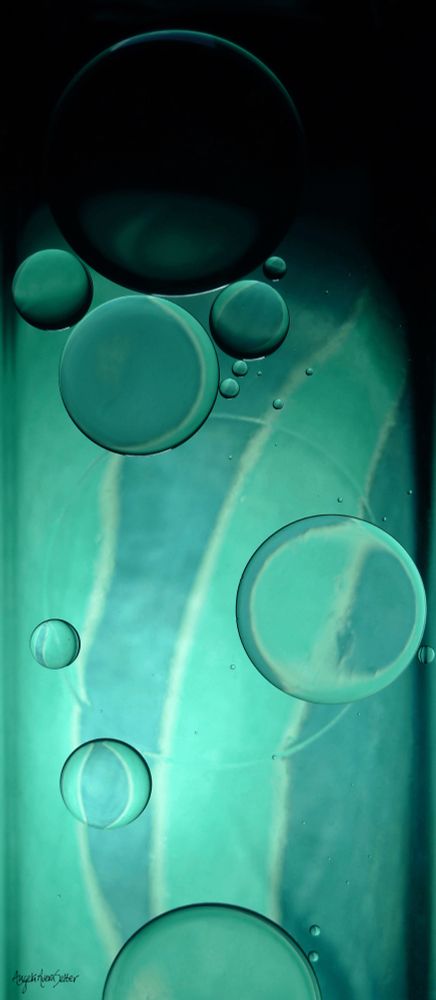 Abstract image, tall vertical arrangement. Hues of aqua waves and stripes fading to deep black at the top of the image. Circular shapes of many sizes float atop the color and light. This was created by floating oil in water in a glass dish atop fabric and illuminated from behind by a light bulb. 