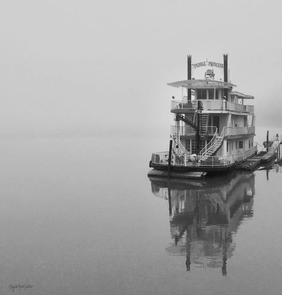 Monochrome image of a paddle-wheel boat docked on the side of a river. The air is very foggy, giving it a feeling of floating with barely a difference between air and water aside from the boat's reflection on the water's surface. Kanawha River, West Virginia, US.