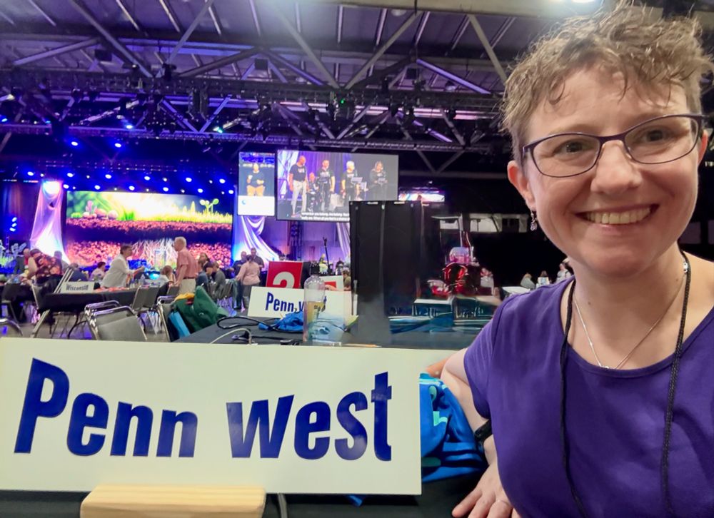 Photo of Nicole with a sign that says Penn West, and in the background is the stage set for opening worship
