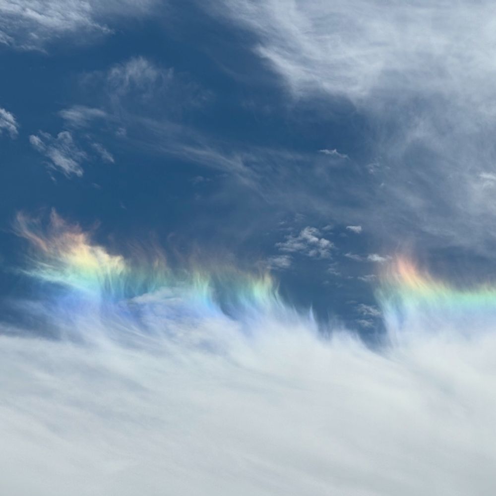 Photo of blue sky with a bank of clouds, the edge of which are wispy and rainbow colored