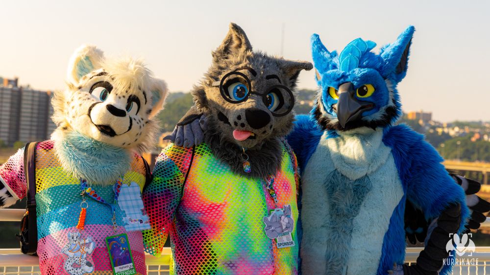 A photo of me in my toony gray wolf fursuit and rainbow net hoodie, Nightwish in his white cheetah fursuit and rainbow net shirt, and Lost in his blue gryphon fursuit, together on the roof of the DLCC at Anthrocon. 