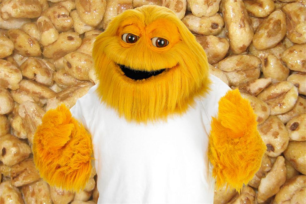 A photo. It's a promotional photo of Honey Monster, the mascot of a brand of cereal in the UK known as Sugar Puffs. He's a big, yellow monster with shaggy fur and brown eyes. He's wearing a plain white t-shirt and can only be seen from the waist up. He's smiling and looking at the camera, with his arms gesturing to himself as if posing for the photographer. The background is a close-up photo of Sugar Puffs cereal.