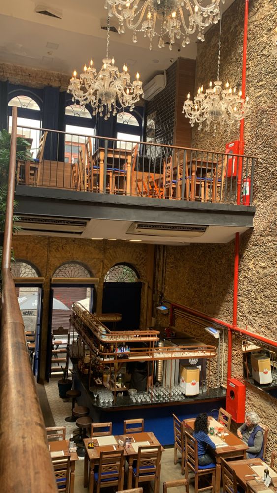 foto de um restaurante de um casarao antigo. a foto está de cima pra baixo e embaixo tem algumas cadeiras e mesas de madeira no teto lustres brancos 