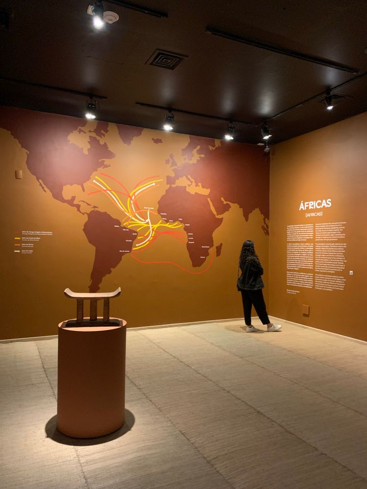foto da exposição ancestral do ccbb. uma foto da sala distantecom o fundo marrom claro(?) de um lado a america do sul a africa uma ponta dos eua e da europa do outro lado da parede está escrito africas em branco e um texto falando sobre a exposição da sala.