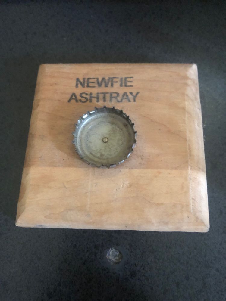 a block of wood with a beer bottle cap nailed to it. The words “Newfie Ashtray” are stamped above the bottle cap. 