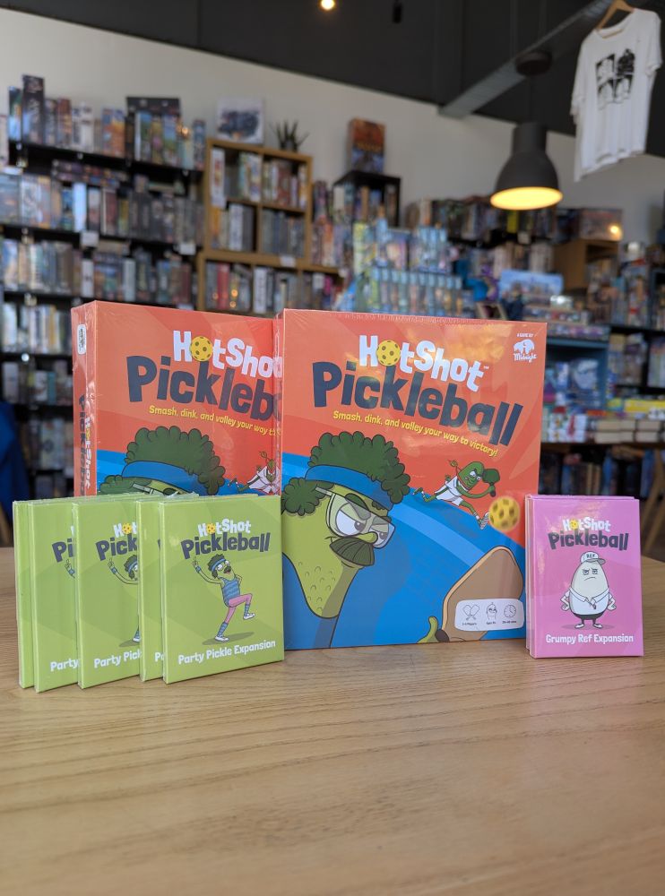 New board game Hotshot Pickleball in the Edinburgh board game shop Ancient Robot Games. The game has illustrations of pickles playing pickleball. Shelves of board games are visible in the background.